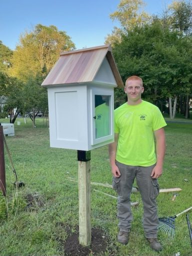 A young man in a bright shirt stands next to a small, wooden library on a post.